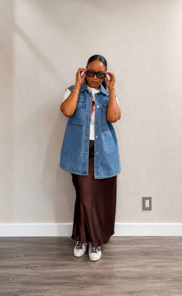 Steffanie Monae wearing brown sunglasses with blueish purple tint, a denim vest, graphic tee, brown satin skirt, and sneakers for an elevated casual outfit